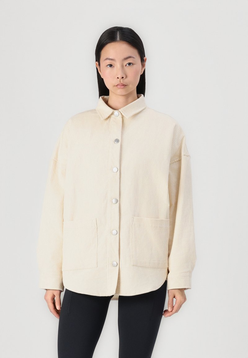 Veste en velours côtelé beige avec col, boutons-pression sur le devant et deux poches plaquées inférieures ; présente une coupe décontractée et une texture côtelée.