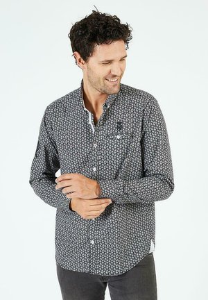 Chemise noire à manches longues avec un motif floral blanc, dotée d'un col. Présente des accents blancs et une petite poche sur la poitrine.