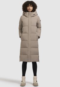 Garšs beiga pufīgais mētelis ar kapuci, ar izšūtu rakstu, sānu kabatām un taisnu siluetu. Valkāts virs melnajām legingām un masīviem zābakiem.