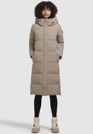 Langer beiger Steppmantel mit Kapuze, der ein gestepptes Design, Seitentaschen und eine gerade Silhouette bietet. Getragen über schwarzen Leggings und klobigen Stiefeln.