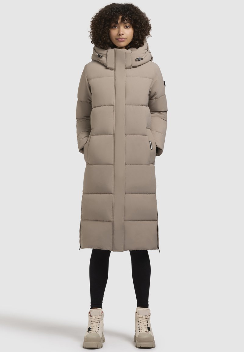 Garšs beiga pufīgais mētelis ar kapuci, ar izšūtu rakstu, sānu kabatām un taisnu siluetu. Valkāts virs melnajām legingām un masīviem zābakiem.