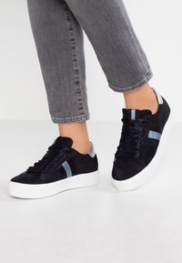 Marinblå mockasneakers med vit sula, utrustade med blå och grå ränder, platta snören och en diskret metallisk detalj på hälen.