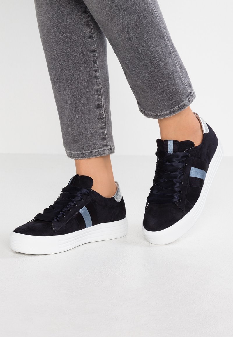 Marinblå mockasneakers med vit sula, utrustade med blå och grå ränder, platta snören och en diskret metallisk detalj på hälen.