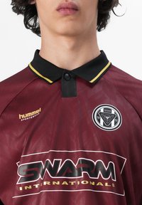 Close-up van een persoon die een bordeauxrode sportpolo draagt met een zwarte kraag, gele bies, "hummel"-logo en tekst "Swarm Internationals" aan de voorkant.