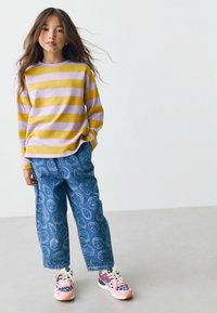 Fille portant un pull rayé jaune et lavande, un jean bleu avec des motifs de smileys, et des baskets en léopard rose, se tenant devant un fond blanc.