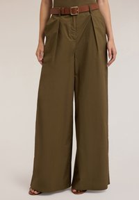 Olijfgroene wijde broek met voorplooien, een bruine riem, gedragen door een persoon die met ontspannen handen aan de zijkant staat.