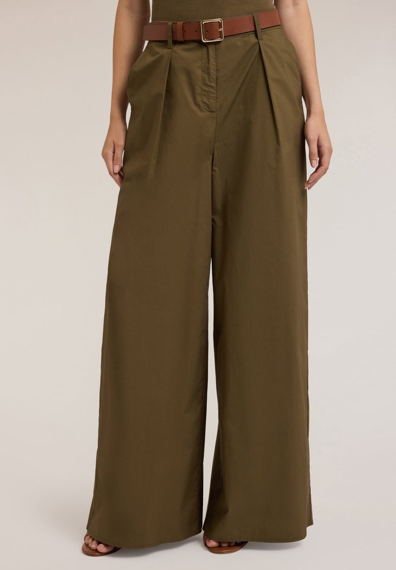 Olijfgroene wijde broek met voorplooien, een bruine riem, gedragen door een persoon die met ontspannen handen aan de zijkant staat.