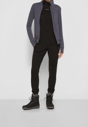 Frau trägt schwarze Hose, schwarzen Rollkragenpullover mit "Icebreaker"-Logo, graue Reißverschlussjacke und schwarze Wanderschuhe, steht vor weißer Wand.