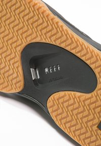 Fekete gumitalp, zigzag mintázattal, világosbarna aljjal és a közepén "REEF" felirattal gravírozott fémcsattal.