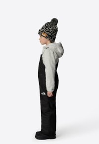 Black waterproof overalls with a fleece-lined hooded jacket, worn with a patterned knit beanie featuring a pom-pom, side profile view.