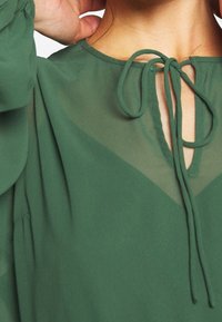 Blusa verde con escote en forma de llave, cierre atado y tejido texturizado. Presenta un ligero brillo y un diseño suave y fluido.