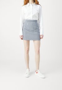 Weißes Hemd mit Knöpfen, karierten marineblauen und weißen Minirock und weiße Sneakers mit roten Akzenten. Der Rock hat eine figurbetonte Silhouette und einen strukturierten Stoff.