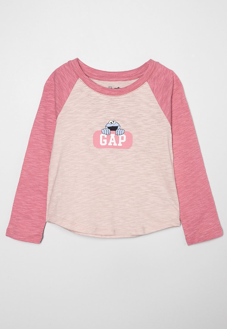 T-shirt à manches longues avec un corps rose clair et des manches raglan rose foncé. Présente un personnage de dessin animé et "GAP" imprimé sur le devant.