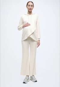 Tenue de maternité comprenant un sweat à capuche crème à boutonnage croisé et un pantalon large assorti. L'ensemble est confectionné dans un tissu doux et texturé.