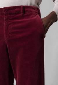 Manšestrové kalhoty barvy burgundské s viditelnou žebrovanou strukturou, s boční kapsou a pasem, nošené nad hnědým topem.