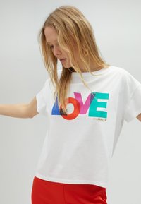 Bílý bavlněný tričko s barevným grafickým motivem "LOVE" v červené, modré a zelené barvě, s textem "by MAITE" ve zlaté. Krátké rukávy a volný střih.