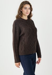 Brązowy sweter z dzianiny z teksturowanym wzorem w warkocze, okrągłym dekoltem oraz ściągaczami przy rękawach i u dołu. Połączony z granatowymi spodniami.