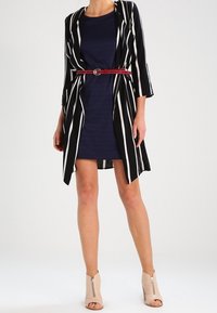 Un long cardigan rayé noir et blanc porté sur une robe navy, cintré à la taille avec une ceinture rouge. Des bottines beige à talons complètent le look.