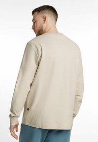 Beige long-sleeve shirt with textured fabric, round neckline, and cuffs. Notable detail includes a small brand tag on the lower side.