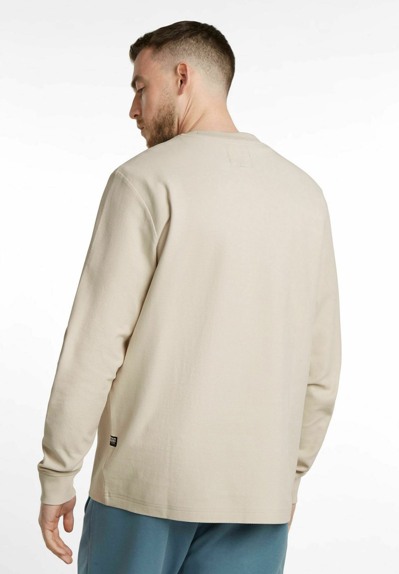 Beige long-sleeve shirt with textured fabric, round neckline, and cuffs. Notable detail includes a small brand tag on the lower side.