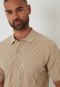 Polo de punto de mangas cortas en color beige con un diseño geométrico texturizado, parte delantera abotonada y cuello clásico con detalles sutiles.
