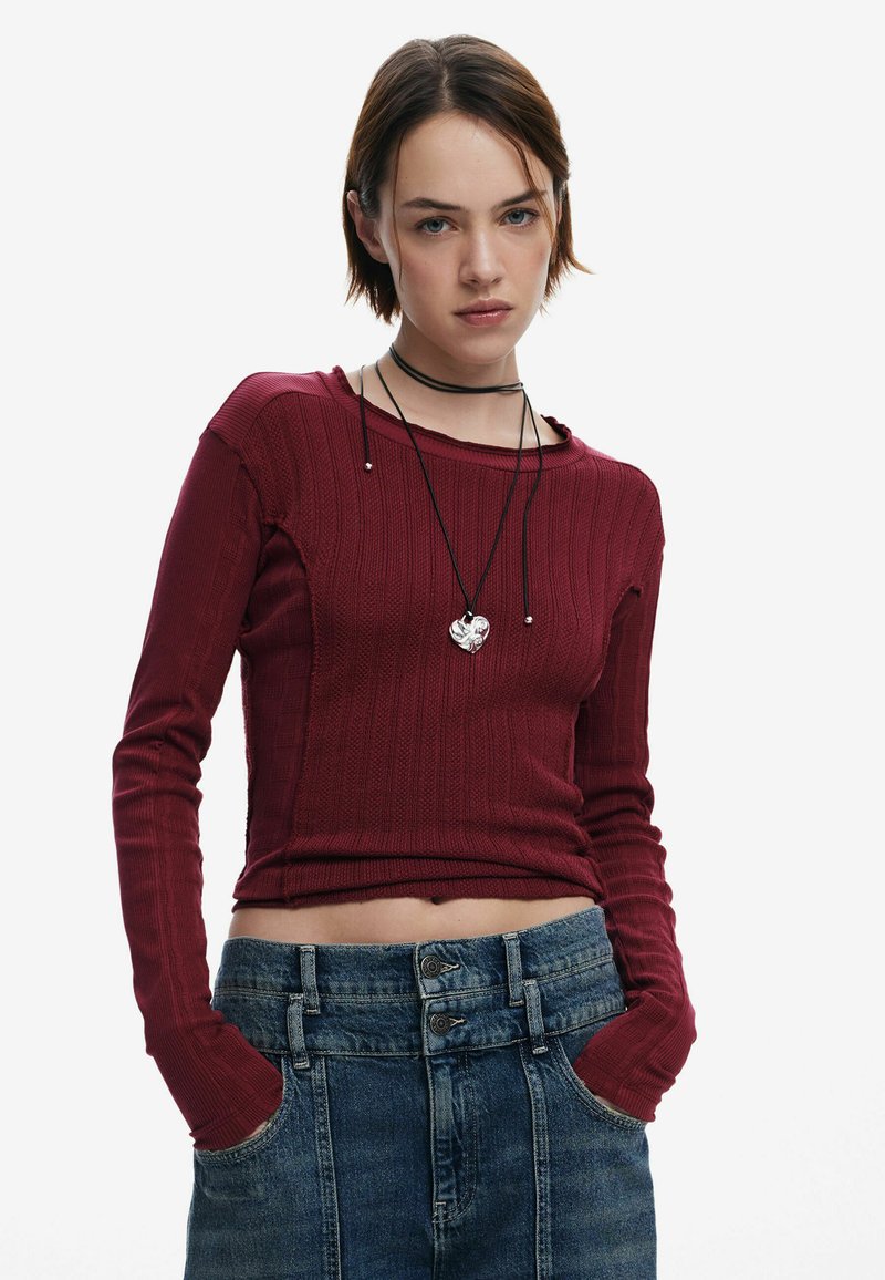 Burgunderfarbene, gerippte Langarmbluse mit einem cropped Design, kombiniert mit hochgeschnittenen, hellblauen Denim-Shorts mit doppelter Knopfleiste und seitlichen Taschen.