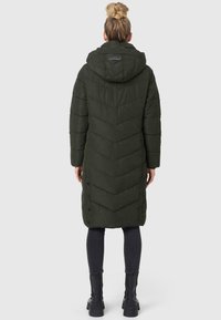 Dunkelgrüner gepolsterter Mantel mit einem Zickzackmuster, Kapuze und seitlichen Knöpfen, der bis zur Mitte der Waden reicht. Kombiniert mit engen schwarzen Hosen und Stiefeletten.