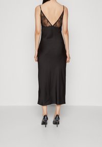 Robe en satin noir avec un dos en V, présentant un panneau en dentelle transparente. Coupe slim, longueur cheville, bretelles fines complètent le design. Chaussures à talons aiguilles visibles.