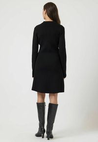 Abito nero a coste con maniche lunghe e una leggera silhouette a trapezio. Abbinato a stivali neri in pelle al ginocchio con dettaglio zip.