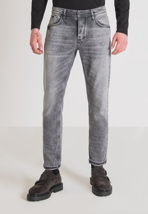 Abgetragene graue ausgebleichte Jeans, kombiniert mit schwarzen Socken und dunkelbraunen Wildlederschuhen, dazu sichtbare Hände und ein schwarzes langärmeliges Oberteil.