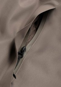 Tejido exterior marrón con un forro suave y texturizado; cremallera expuesta y detalles de costura visibles. Presenta un diseño elegante y ajustado.