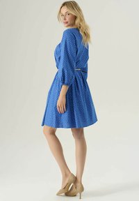 Abito blu a pois con maniche a tre quarti e vita aderente, caratterizzato da un piccolo dettaglio dorato sul retro e una gonna svasata.