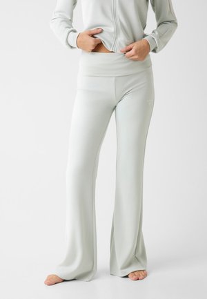 Osoba ubrana w jasnoszare, wysokie spodnie dzwony i pasującą kurtkę na zamek, stojąca boso na jednolitym białym tle.