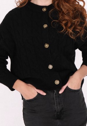 Femme aux longs cheveux bouclés portant un cardigan noir boutonné et un jean en denim foncé, les mains dans les poches avant.