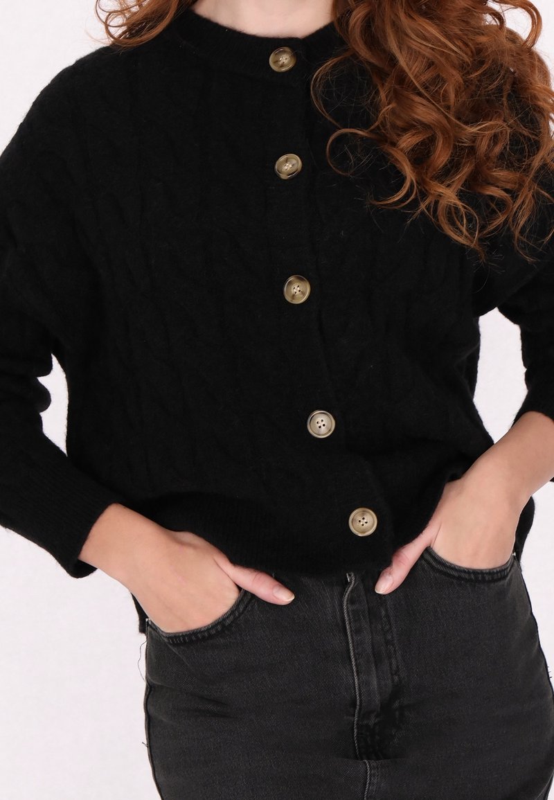 Femme aux longs cheveux bouclés portant un cardigan noir boutonné et un jean en denim foncé, les mains dans les poches avant.