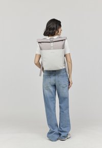 Femme portant une chemise blanche et un jean bleu ample, portant un sac à dos beige et gris à rabat, de dos contre un fond gris uni.