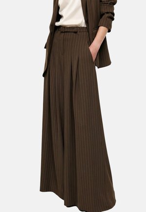 Pantaloni marroni a gamba larga con righe sottili e pieghe frontali, abbinati a un blazer coordinato e una maglietta bianca, con la mano in tasca.