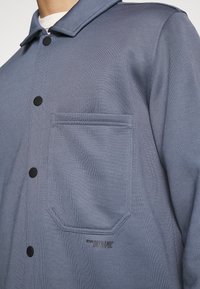 Camisa azul de botones hecha de tela texturizada, con un bolsillo en el pecho y botones negros. El logo "DRYNAMIC" es visible cerca del bolsillo.