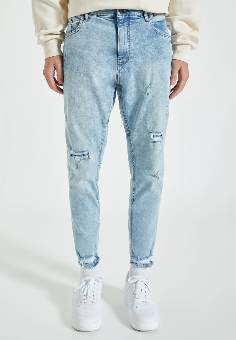 Jean bleu clair déchiré avec des ourlets effilochés porté avec des chaussettes blanches et des baskets blanches, associé à un haut à manches longues couleur crème.