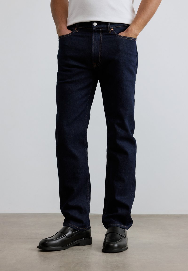 Σκούρο μπλε τζιν με ευθεία γραμμή, διαθέτει εμπρόσθιες τσέπες, διακριτικές ραφές και κλασικό κούμπωμα με κουμπί. Συνδυάζεται με μαύρα μοκασίνια.