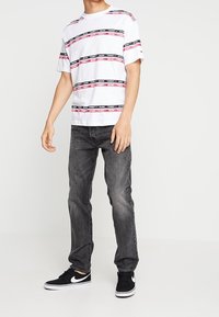 Vit T-shirt med horisontella röda och svarta ränder, texten "TOMMY JEANS", kombinerad med mörkgrå jeans och svarta sneakers.