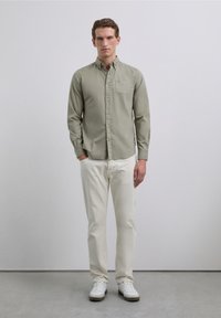 Uomo che indossa una camicia verde chiaro con bottoni, pantaloni bianco sporco e sneakers bianche, in piedi contro uno sfondo grigio chiaro uniforme.