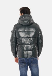 Giacca a vento verde scuro con finitura lucida, cappuccio e sezioni a trapunta orizzontale, abbinata a pantaloni jeans blu.