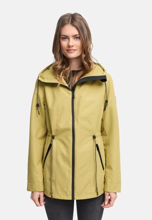 Jeune femme aux cheveux ondulés portant une veste à capuche vert clair avec des détails de fermeture éclair et de poche noirs sur un fond uni.