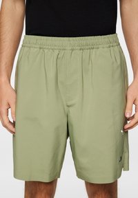 Vaaleanvihreät shortsit, jotka on valmistettu sileästä kankaasta, ja niissä on joustava vyötärö sekä sivutaskut. Sivussa on minimalinen logotyyppi.