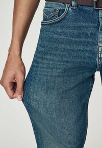 Blugi din denim cu o nuanță medie de albastru, cu o croială clasică; dispune de buzunare, cusături contrastante și un material texturat cu o ușoară decolorare.