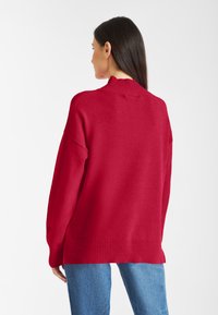 Rode trui met een hoge, geribbelde kraag, drop shoulders en een ontspannen pasvorm. De stof lijkt zacht en wordt gecombineerd met lichtblauwe jeans.