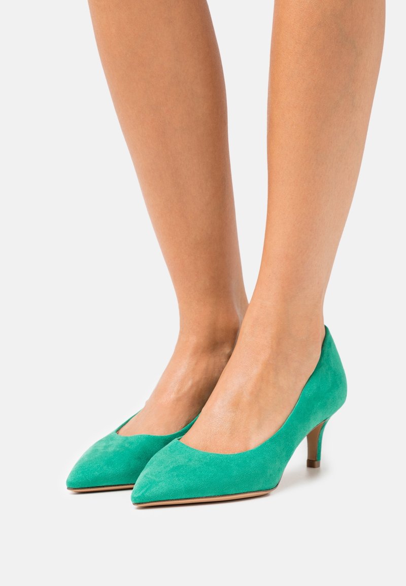 Jambes d’une femme portant des chaussures en daim bleu sarcelle à bout pointu et talons kitten sur un fond clair uni.