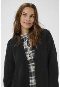 Zwarte, gezellige cardigan met een gestructureerd breipatroon, gedragen over een grijs-wit geruit shirt met een kraag en knoopdetails.