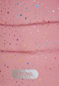 Rosa fluffig jacka med en slät textur, som har flerfärgade glittriga prickar och zigzag-logotyp i silver på en vit bakgrund.